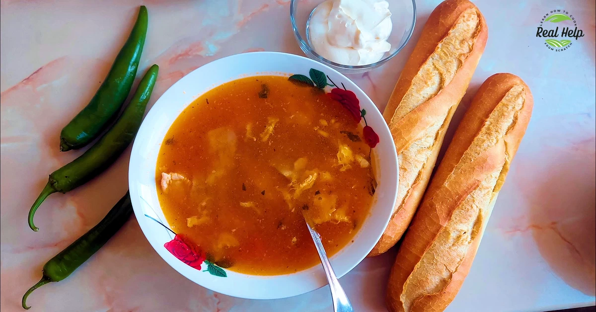 A Bowl of Chicken Borscht Soup Garnished With Fresh Herbs, Accompanied by Sliced Peppers, a Piece of Crusty Bread, and a Dollop of Sour Cream on the Side.